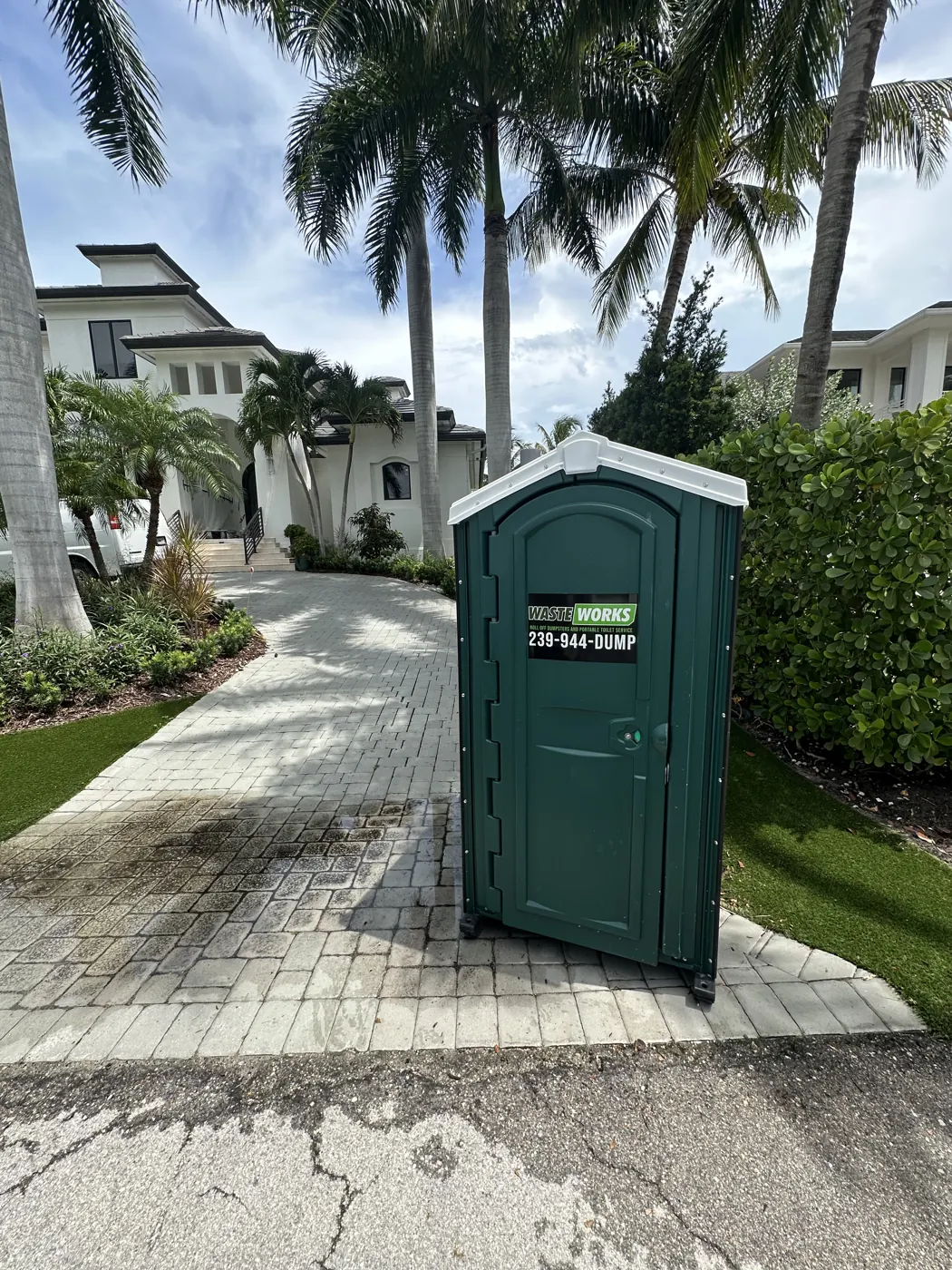 A Waste Works portable toilet beside a palm-lined driveway